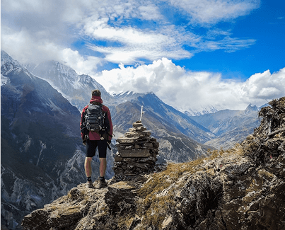 Backpacker standing on a rocky mountain peak with a scenic view of surrounding mountains and a bright blue sky.