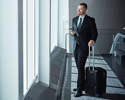 Man in business attire with a suitcase standing at an airport terminal window, looking outside.