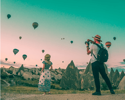 Couple taking photos of hot air balloons in a scenic outdoor location with mountains in the background.
