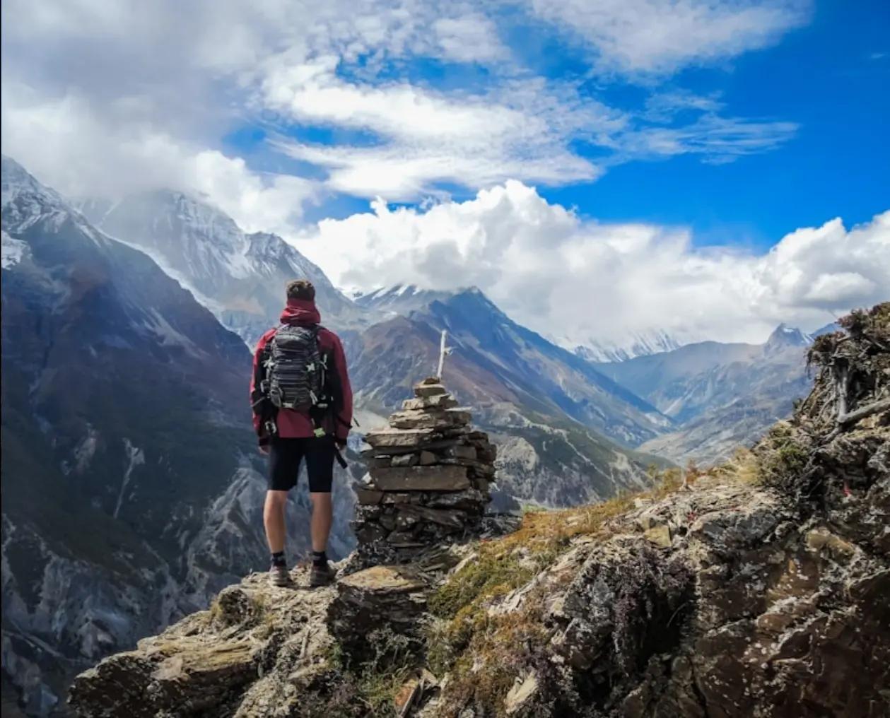 Backpacker standing on a rocky mountain peak with a scenic view of surrounding mountains and a bright blue sky.