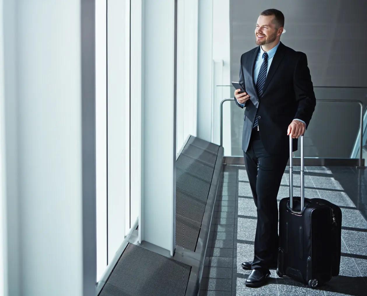 Man in business attire with a suitcase standing at an airport terminal window, looking outside.