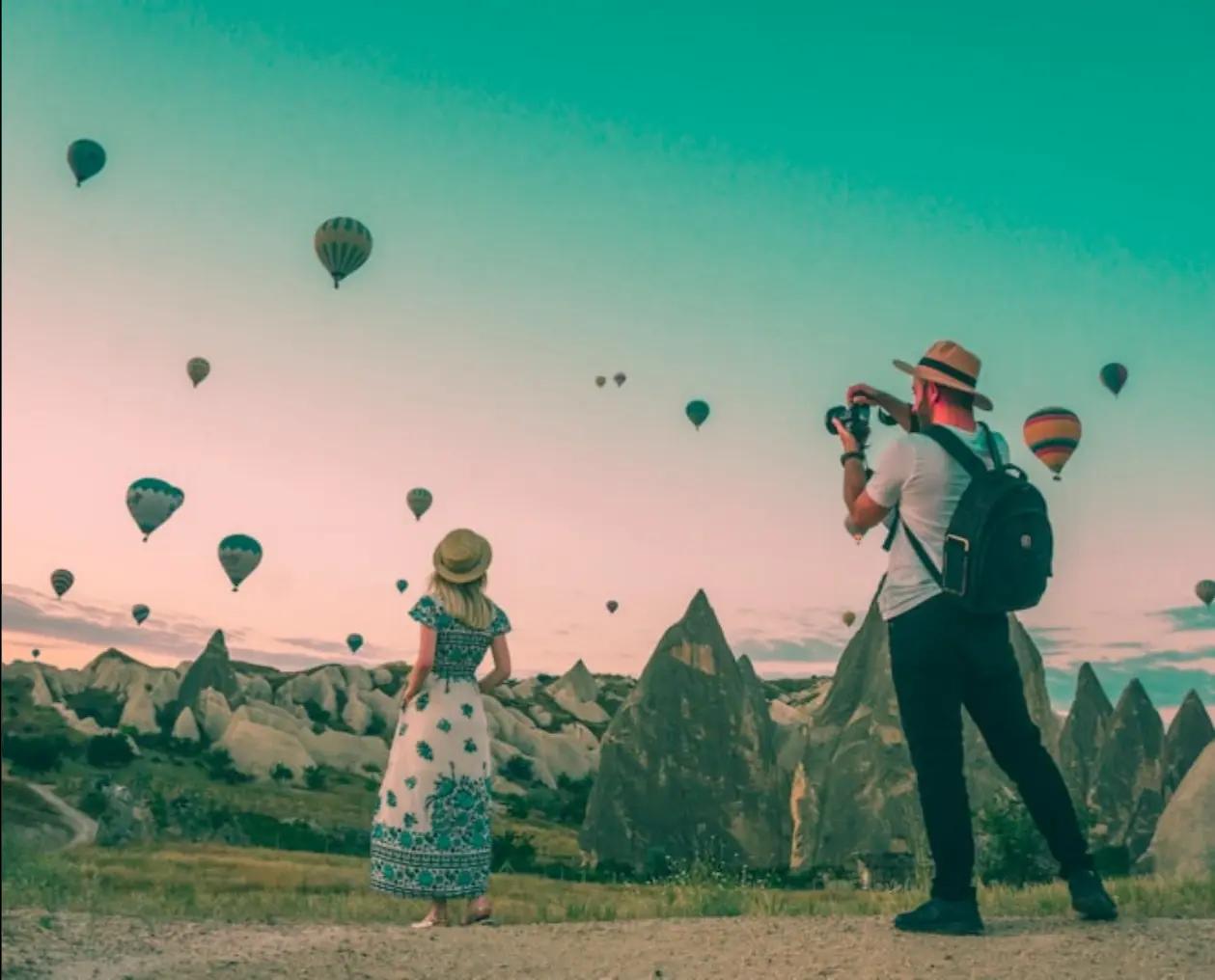 Couple taking photos of hot air balloons in a scenic outdoor location with mountains in the background.